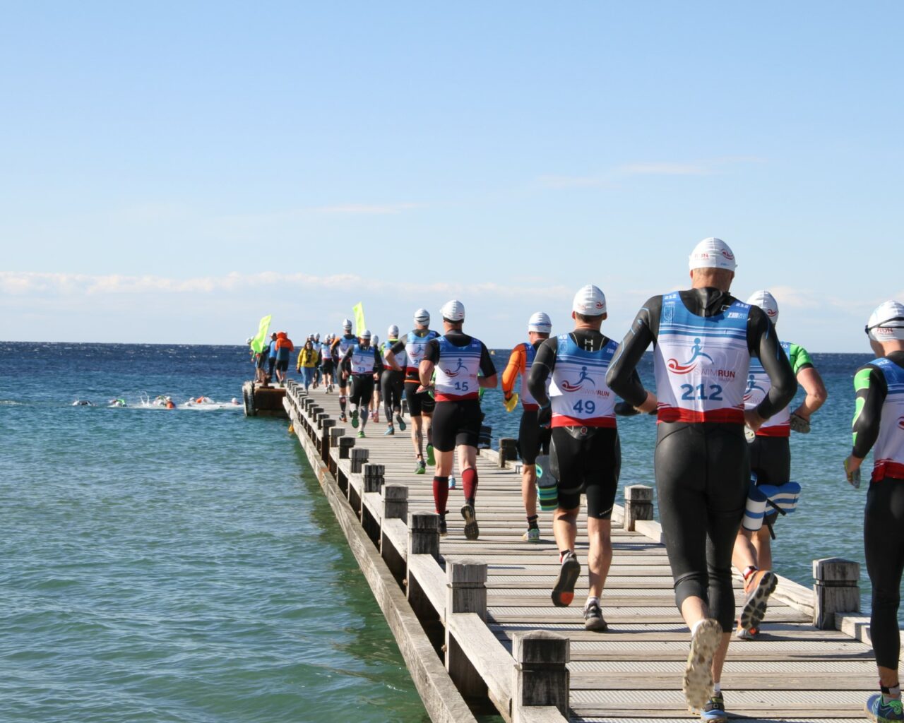 Coureurs en train de se diriger vers la mer depuis lon ponton de la plage du Débarquement pour se lancer à l'eau et continuer leur course. Ils ont tous des combinaisons et leur dossard.