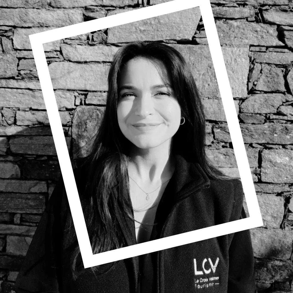 Charmante Community Manager de l'Office de Tourisme qui sourit devant un mur de pierre avec le soleil dans le visage, avec une veste aux couleurs de l'entreprise