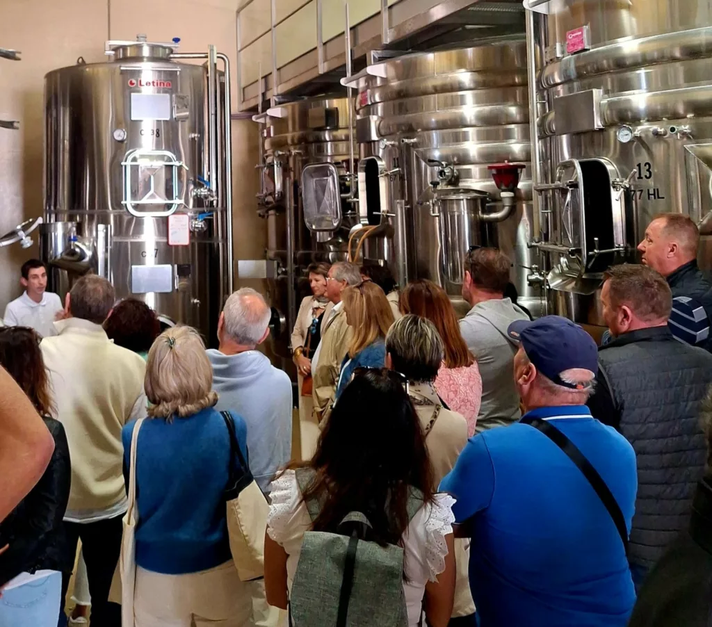 Une foule est en train d'écouter ce que le caviste leur explique à propos de la fabrication du vin. Ils sont tous dans la cave d'un domaine, au milieu des cuves en inox.