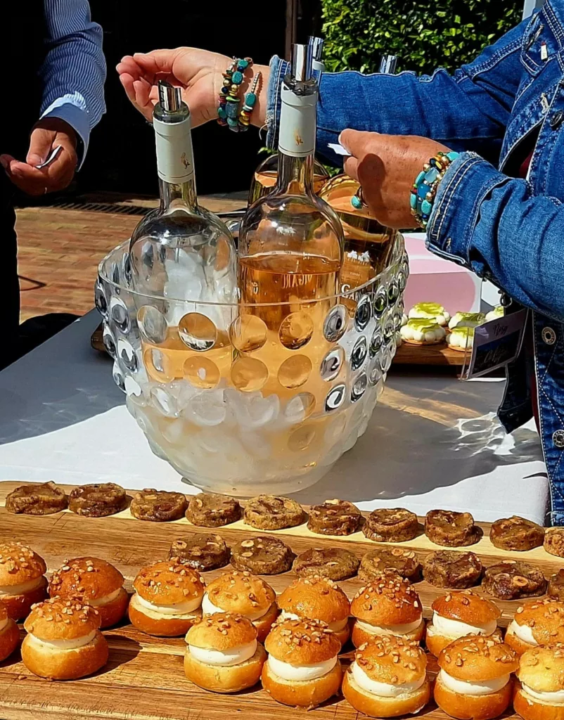 Une dame récupère le ticket d'un monsieur pour le servir, sur la table il y a des bouteilles de vin dans une vasque avec de l'eau et des glaçons; sur une planche en bois il y a des biscuits aux noisettes ainsi que des choux farcis avec de la crème.