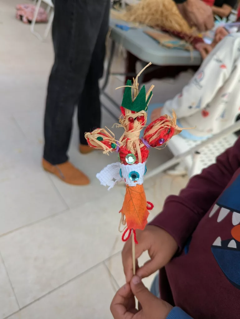 Un enfant présente un jouet qu'il a fabriqué lui même avec du fils, de la paille, des feuilles, du tissus et des brillants au bout d'un pic en bois. Il y a d'autres enfants autour qui sont installés à des tables pour faire la même chose.