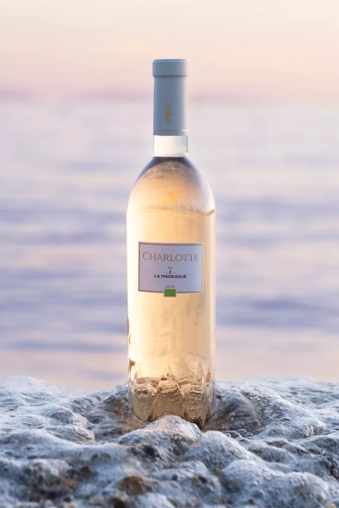 ©Melanie Trupia Garcia Cuvée Charlotte du domaine de la madrague. La bouteille est posée sur le sable et une vague vient taper dessus. C'est la fin de journée car le couleur présument un coucher de soleil.